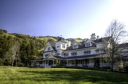 A large white house with black roofing in front of green hills and forests.