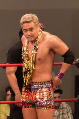 Kazuchika Okada, a Japanese man with blonde hair, wears a golden garland as well as red and gold trunks while standing in a professional wrestling ring