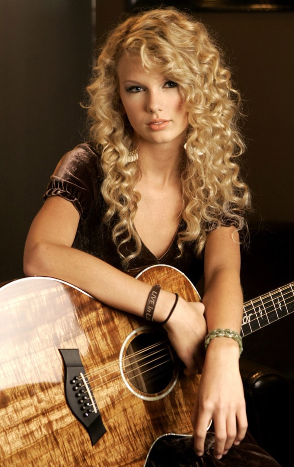 A young female with curly blond hair faces down at an acoustic guitar made of koa wood while a large microphone is placed close to her. She is wearing patterned, red dress.